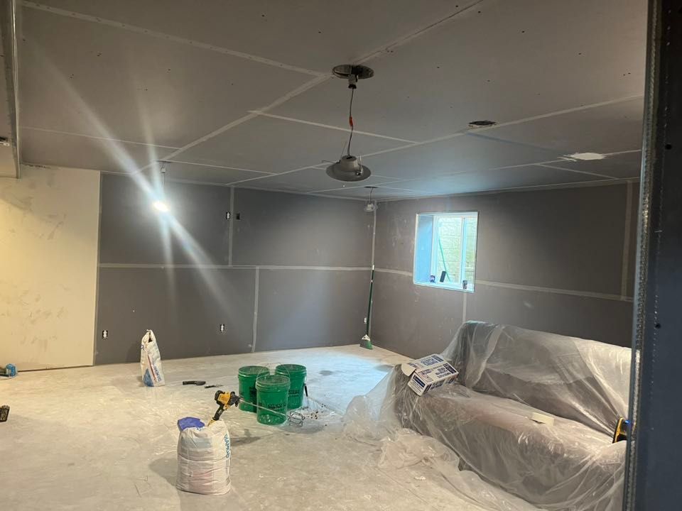 A room under renovation with drywall installed on walls and ceiling, a covered couch, and construction buckets on the floor.