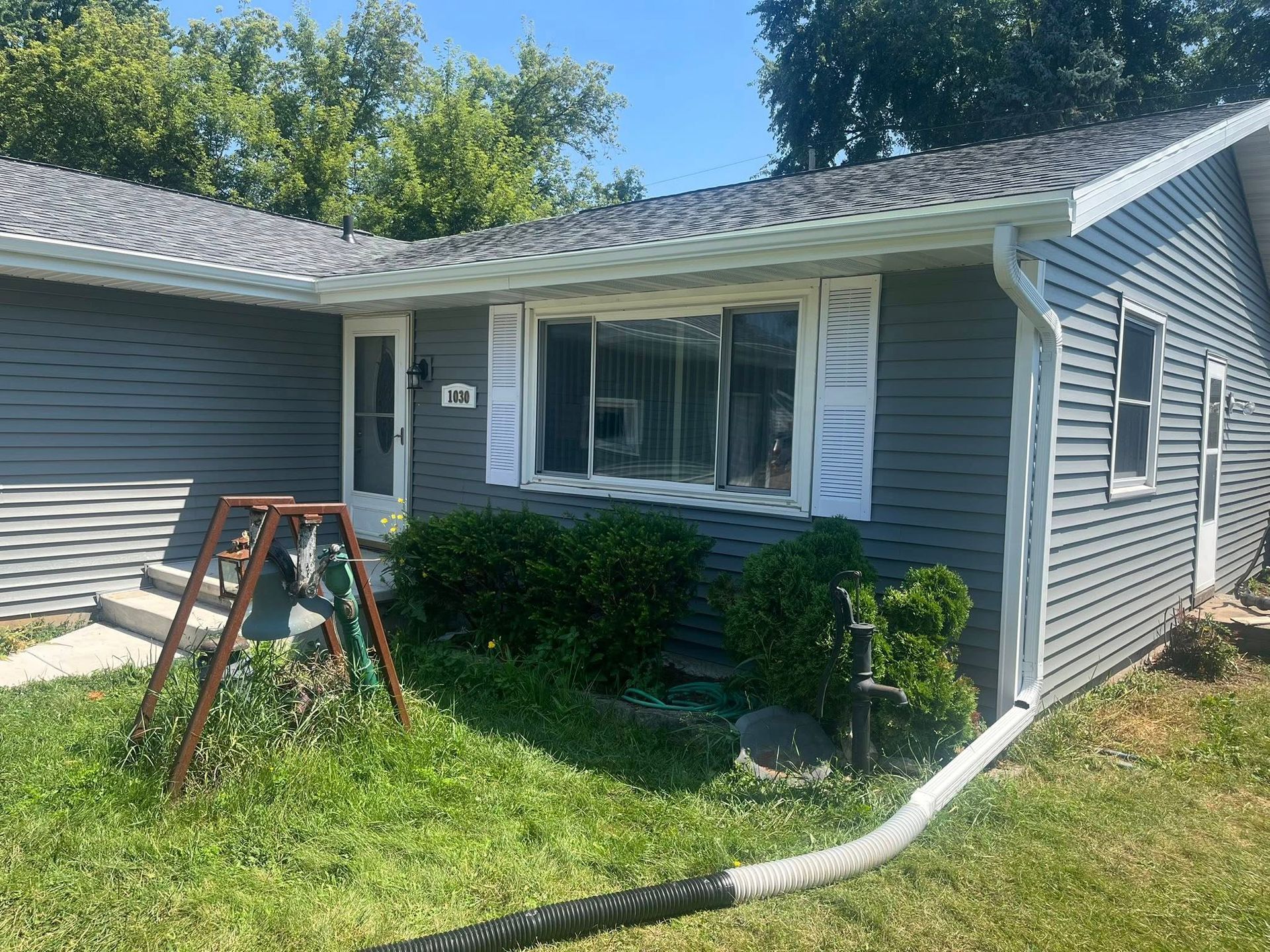 A one-story gray house with white trim and shutters, featuring a small lawn and an extended white gutter downspout.