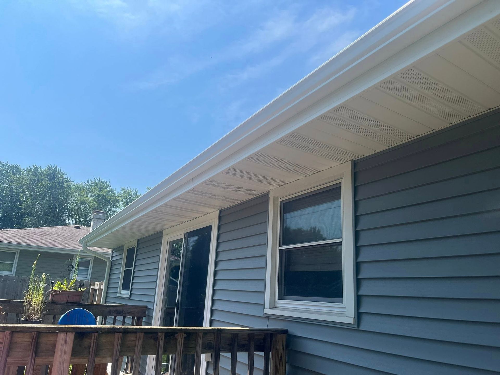 A low-angle view of a gray-sided house exterior with a white soffit, gutter, and window under a bright blue sky.