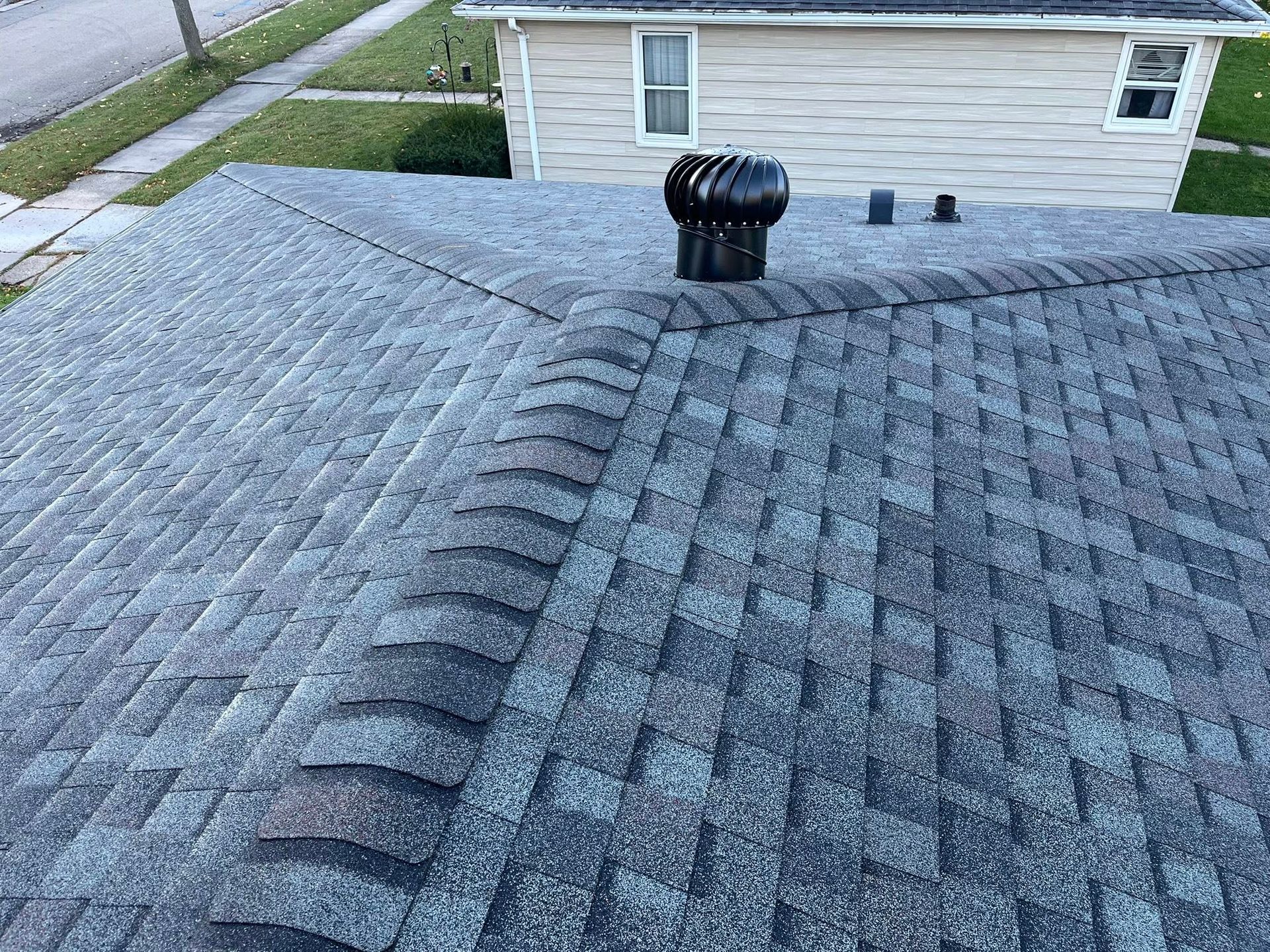 An elevated view of a gray asphalt shingle roof with a central ridge vent and a black rotating roof turbine ventilator.