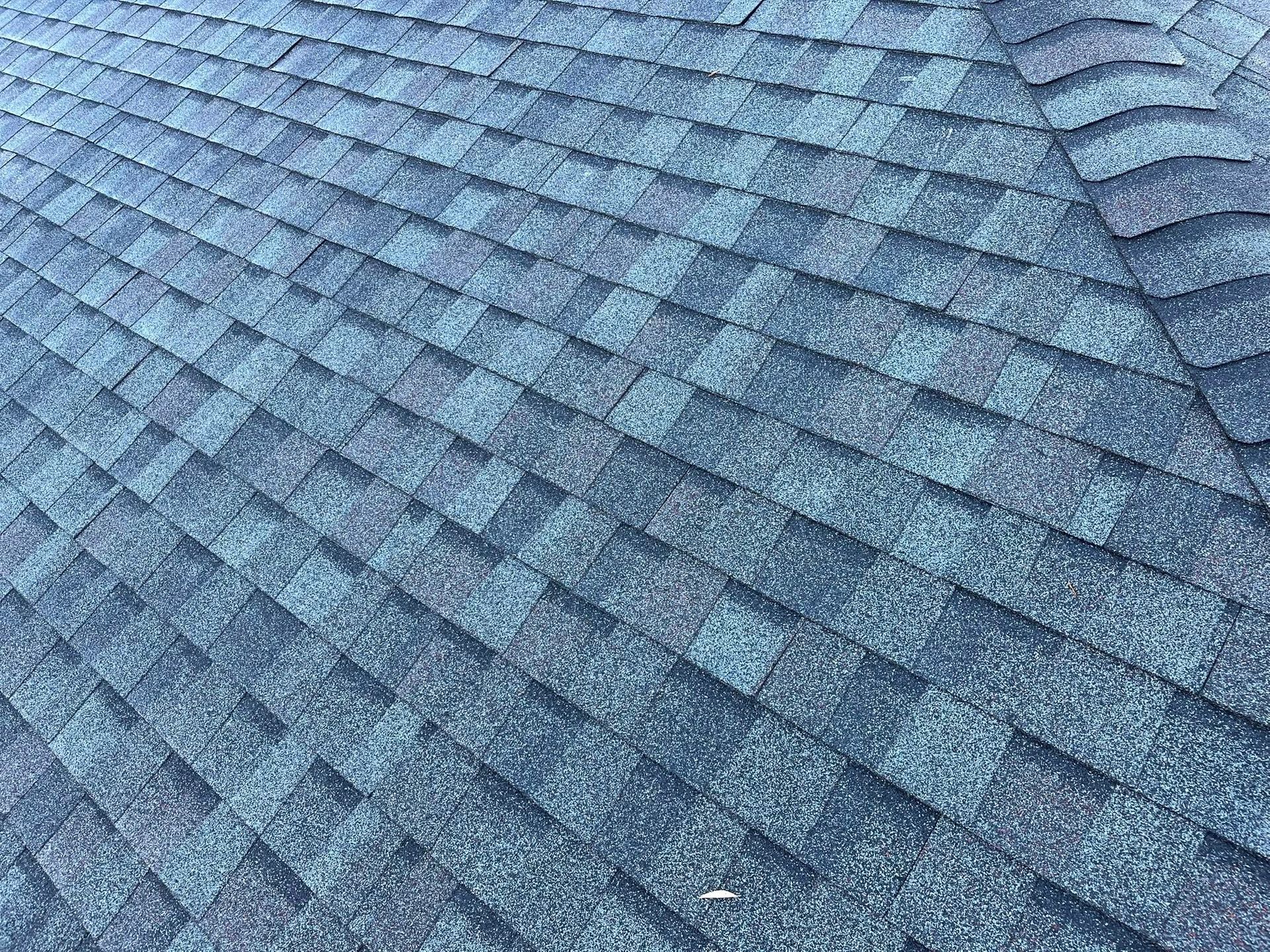 A close-up view of dark grey and blue asphalt shingles on a roof, showing the layered texture and pattern.
