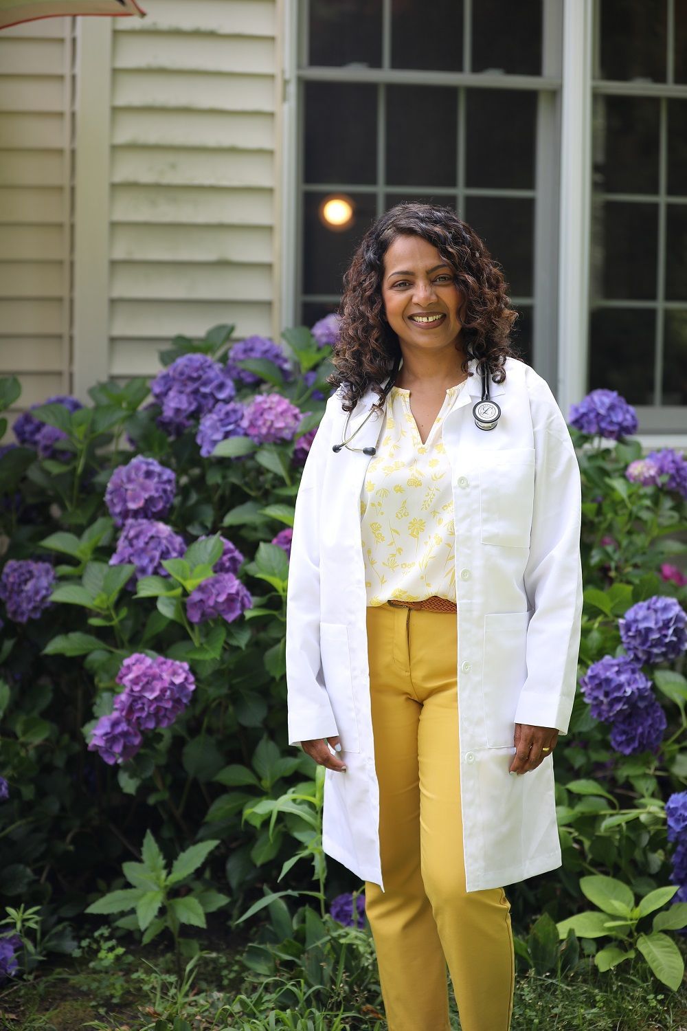 Woman in white coat and mustard pants standing in front of purple hydrangeas and a house window.