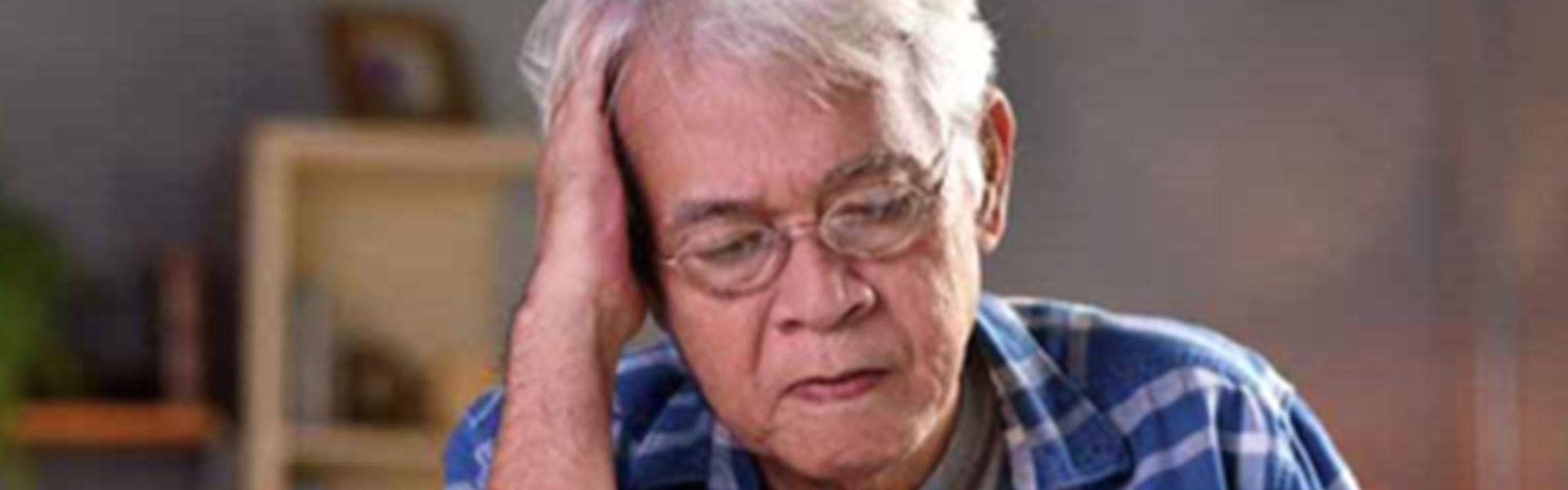 Older person in a blue plaid shirt holding their head with a pained expression indoors
