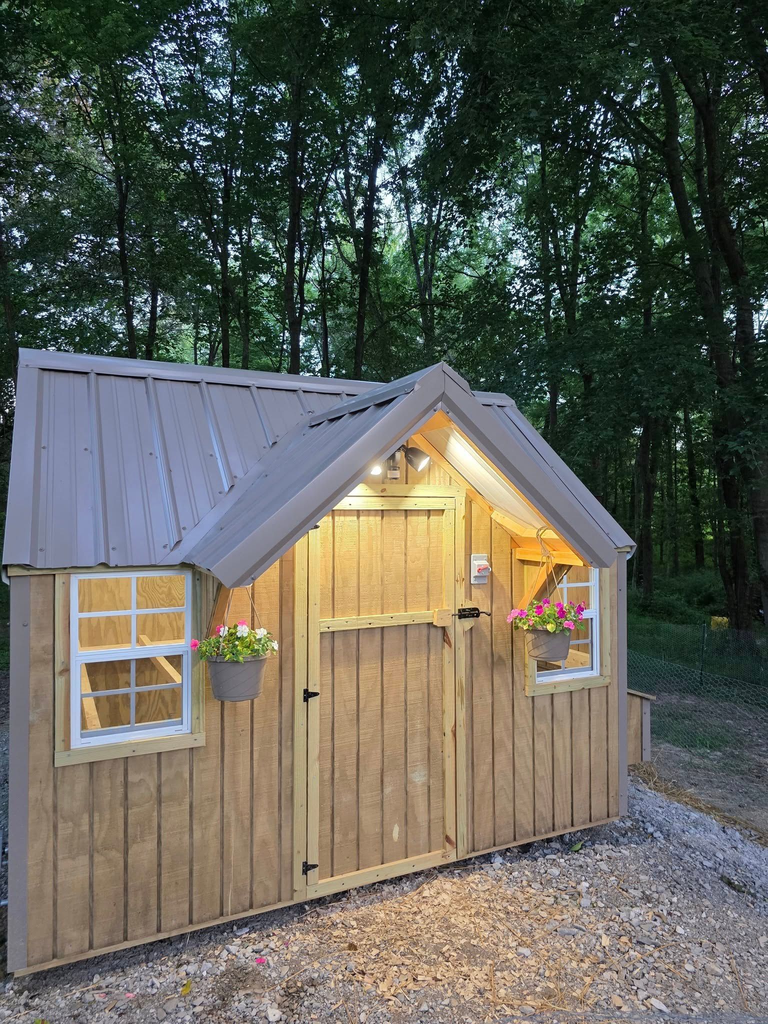 Wooden shed with metal roof, windows, and hanging flower baskets, lit at night in a wooded area.
