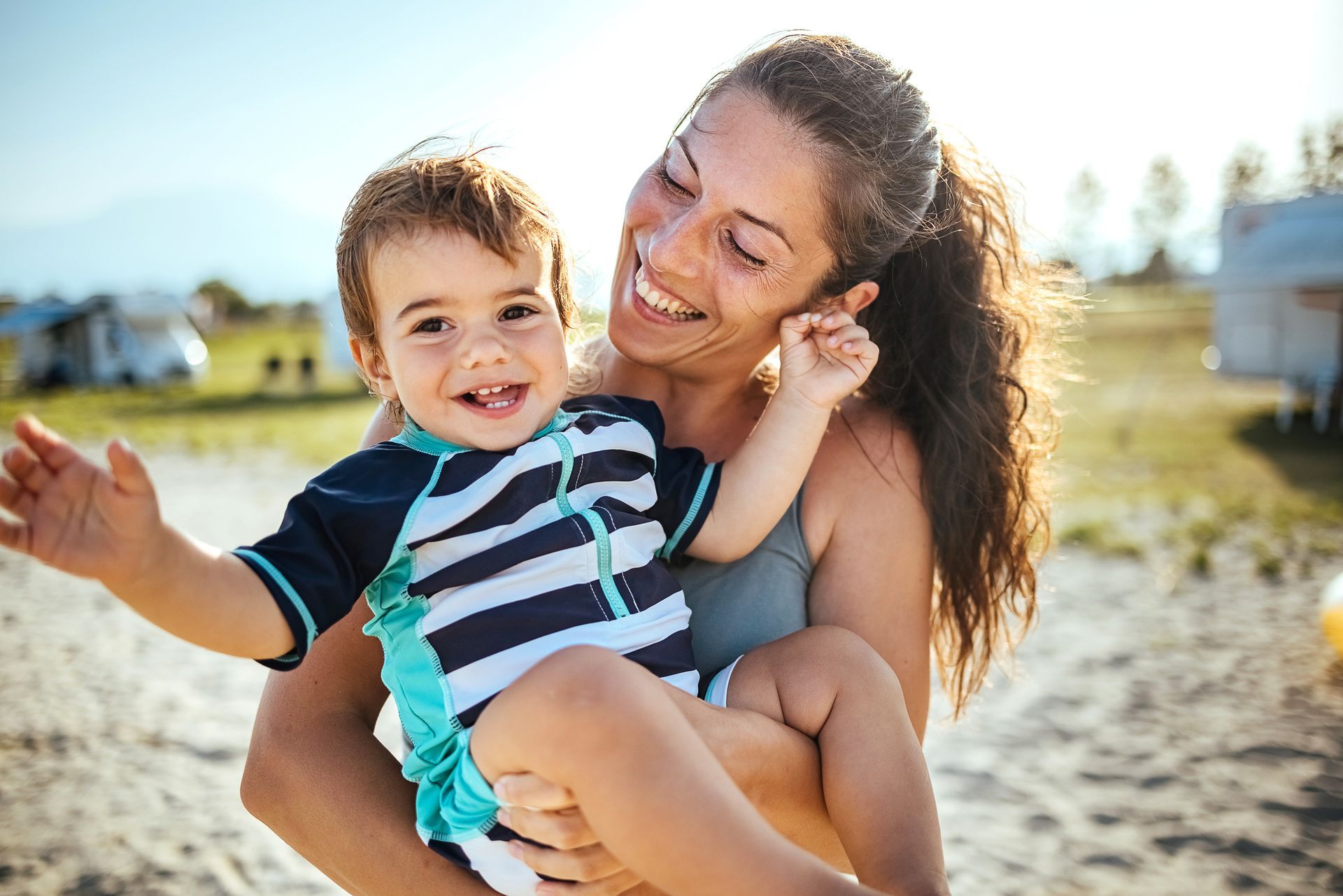 A mother is holding her son in her arms. They are smiling and she is standing outside with sand and grass.