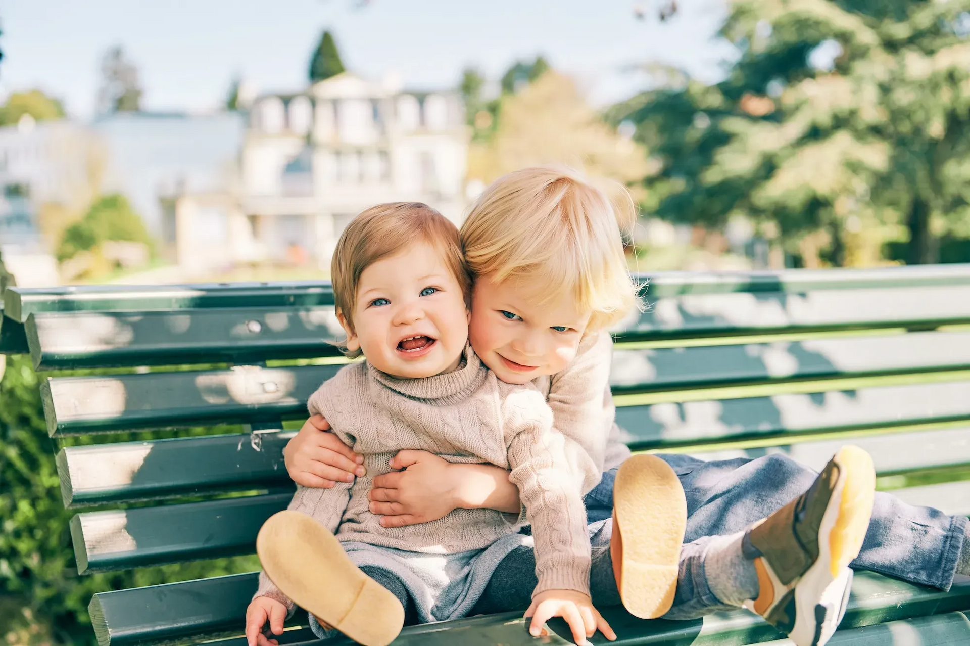 Two young children sitting on a park bench with trees in the background.