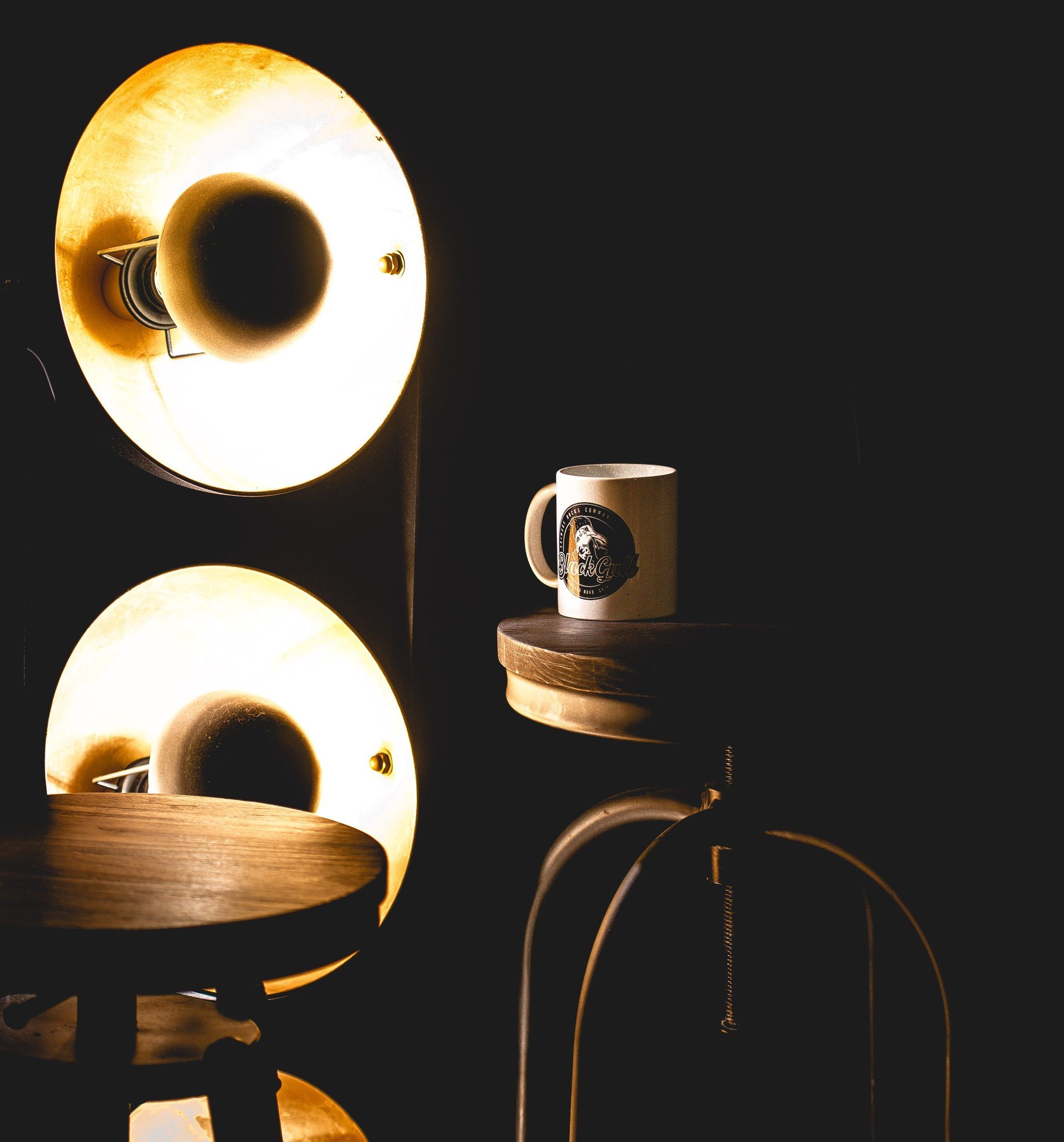 Two lit lamps next to a stool and a mug on a small shelf against a dark wall.