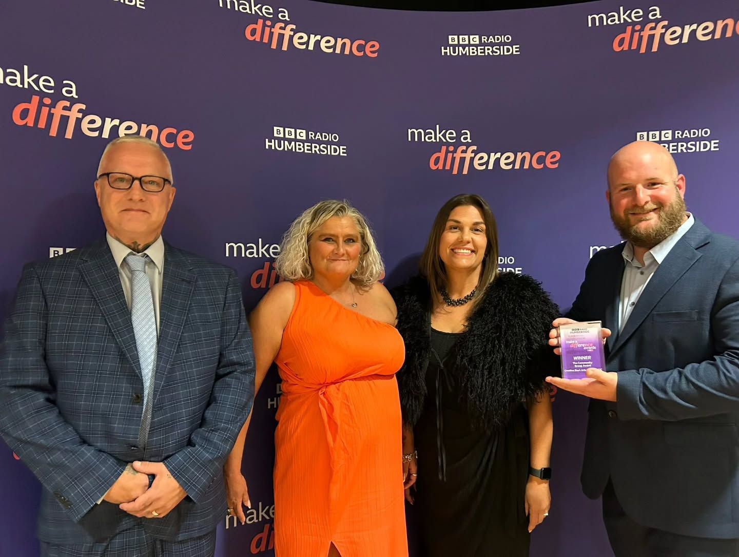 Four people at an event, holding an award. Purple backdrop with