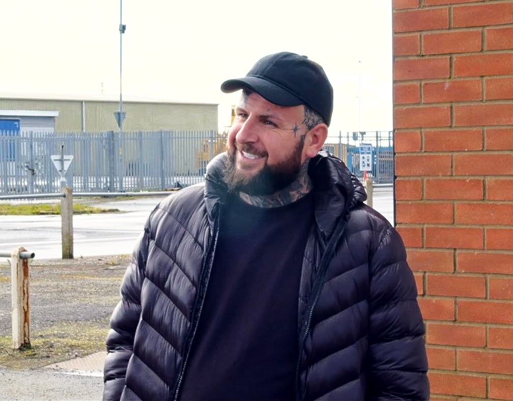 Lisa working on the 'Fanzone' Mural at Blundell Park in Grimsby. Man with beard and tattoos wearing black cap and jacket, smiling near a brick wall and fence.