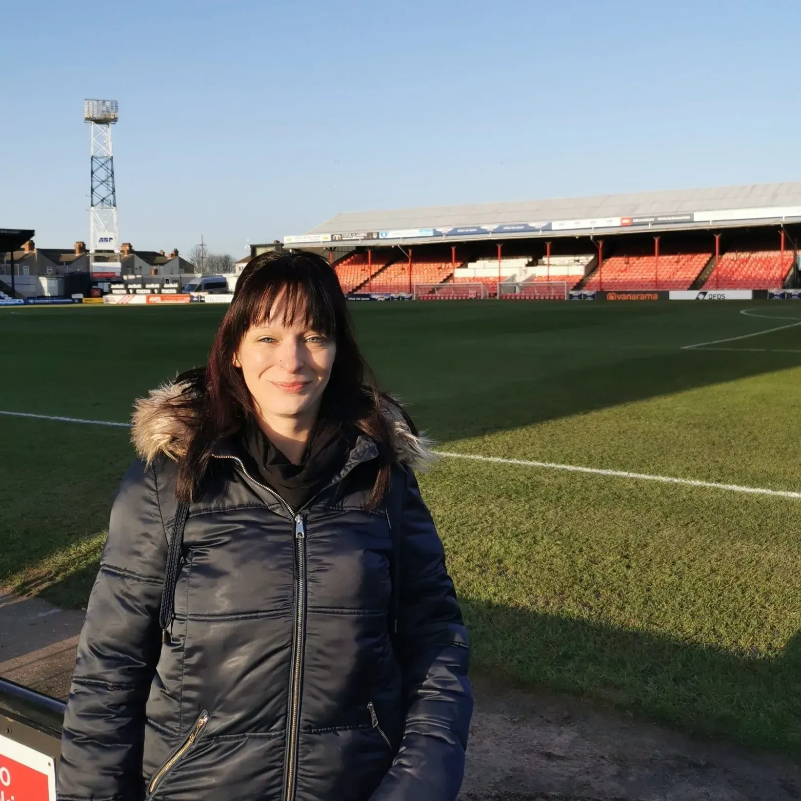 Woman standing in front of a soccer stadium; sunny day. She is wearing a dark coat. Stadium has red seating.