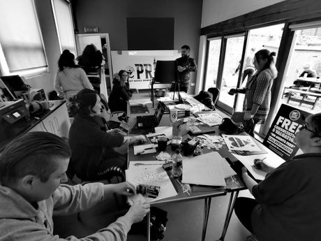 People working at a table, some holding signs, in a room with a large window and sign on the wall.