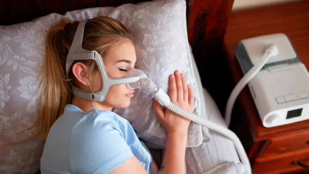 Woman using a CPAP mask while lying in bed beside a bedside machine