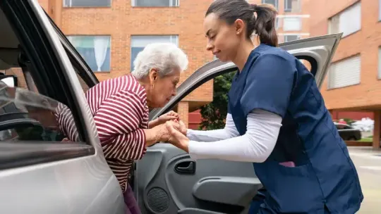 Caregiver helping an elderly woman into a car outside a building