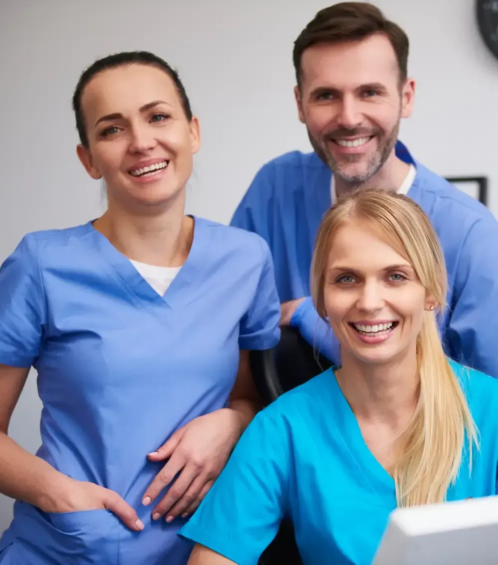 Three healthcare workers in blue scrubs smiling in a clinical office