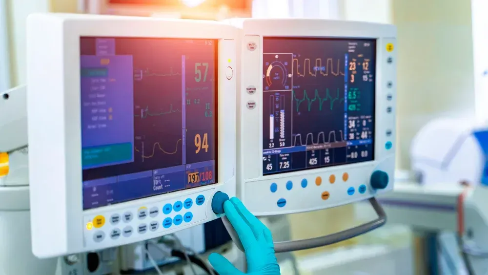 Medical monitor screens with vital signs in a hospital setting, one touched by a gloved hand