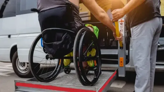Person in wheelchair boarding a van using a portable ramp, with two helpers nearby