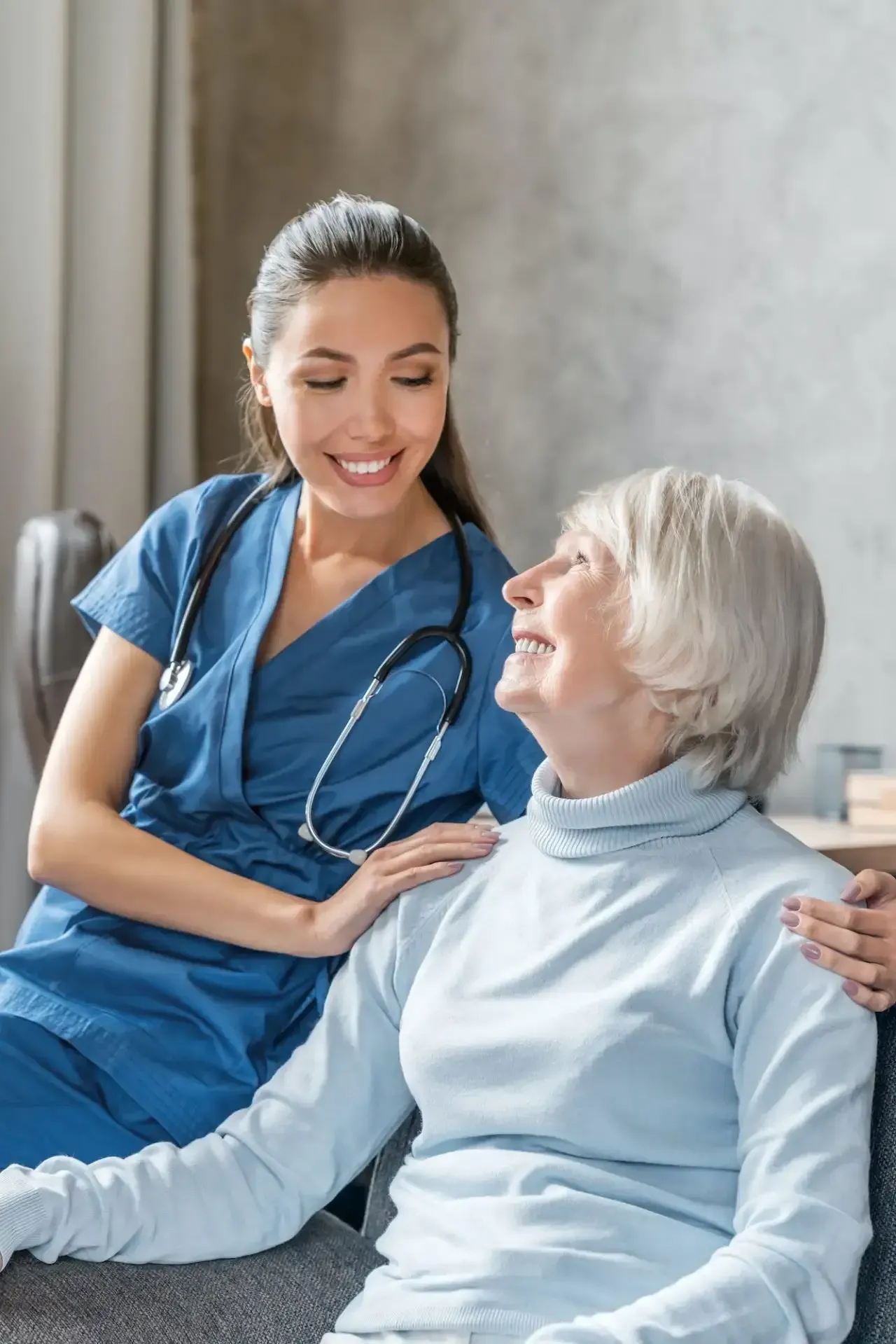 Nurse in blue scrubs smiling and talking with an elderly patient in a hospital room