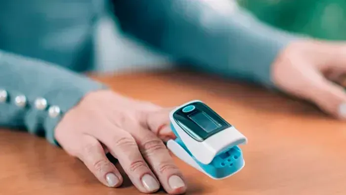 Hand wearing a blue pulse oximeter on a wooden table, with a blurred person in the background