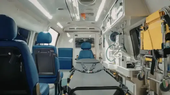 Inside an ambulance, a stretcher faces blue seats and medical equipment along the right wall.
