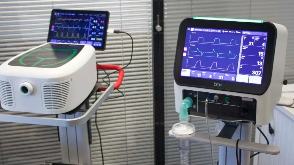 Two medical monitors on carts displaying vital signs, one with a ventilator-like device attached