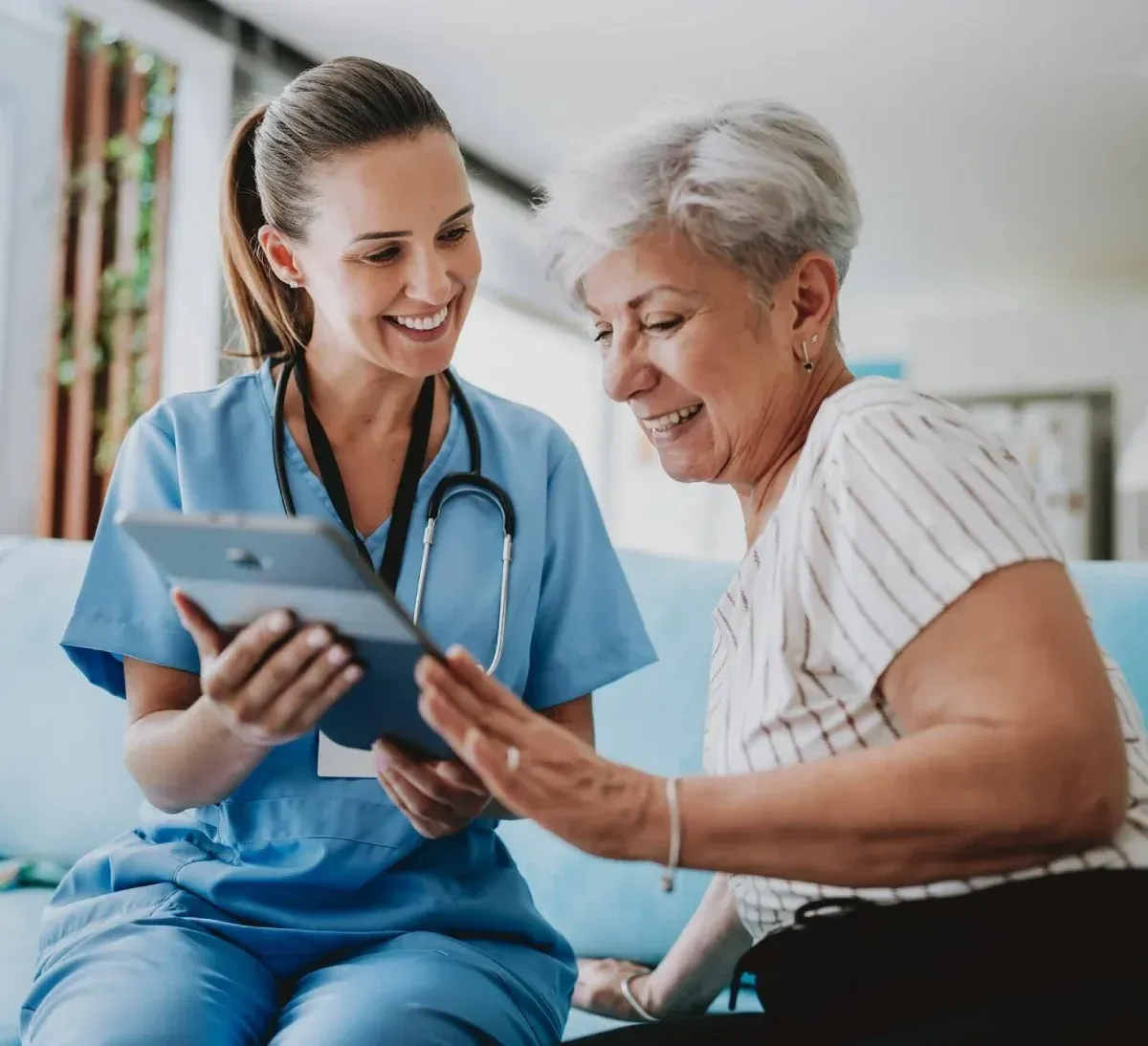 Nurse in blue scrubs showing a tablet to a smiling patient in a bright clinic room