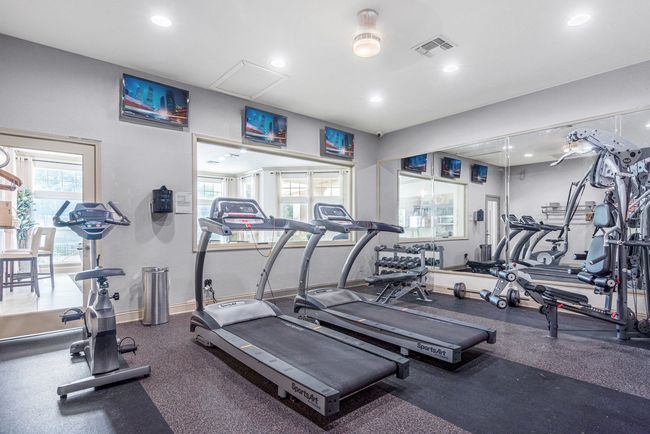 Gym interior with cardio equipment and weight machines. Windows, mirrors, and TVs visible.