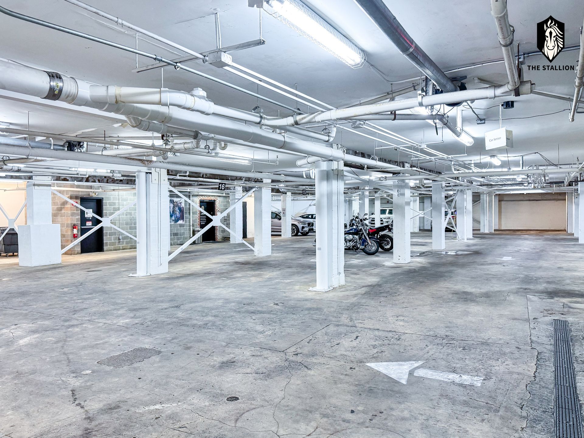 Empty underground parking garage with white pillars, exposed pipes, and concrete floor.