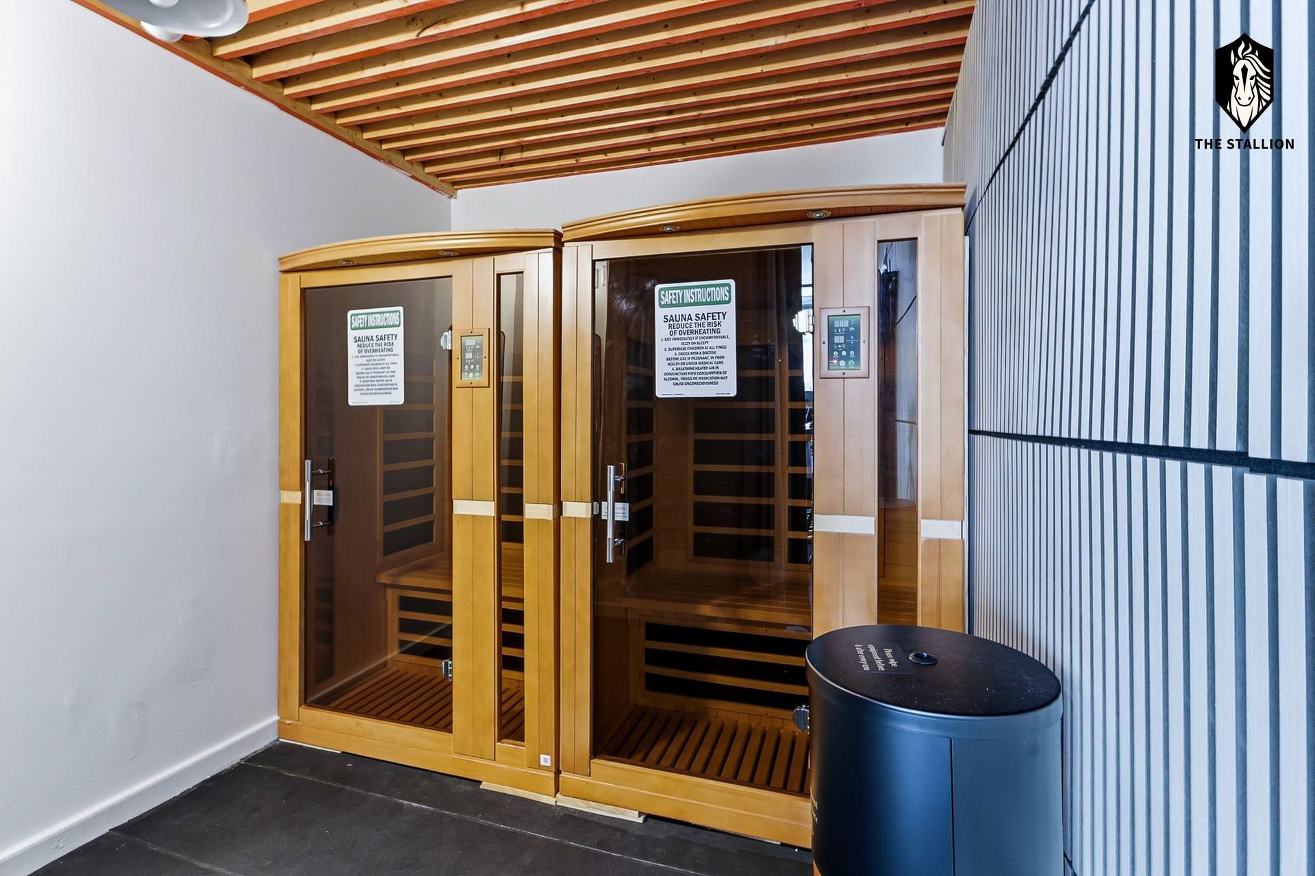 Small wooden sauna cabin with glass doors in a hallway, beside a blue wall and trash can.