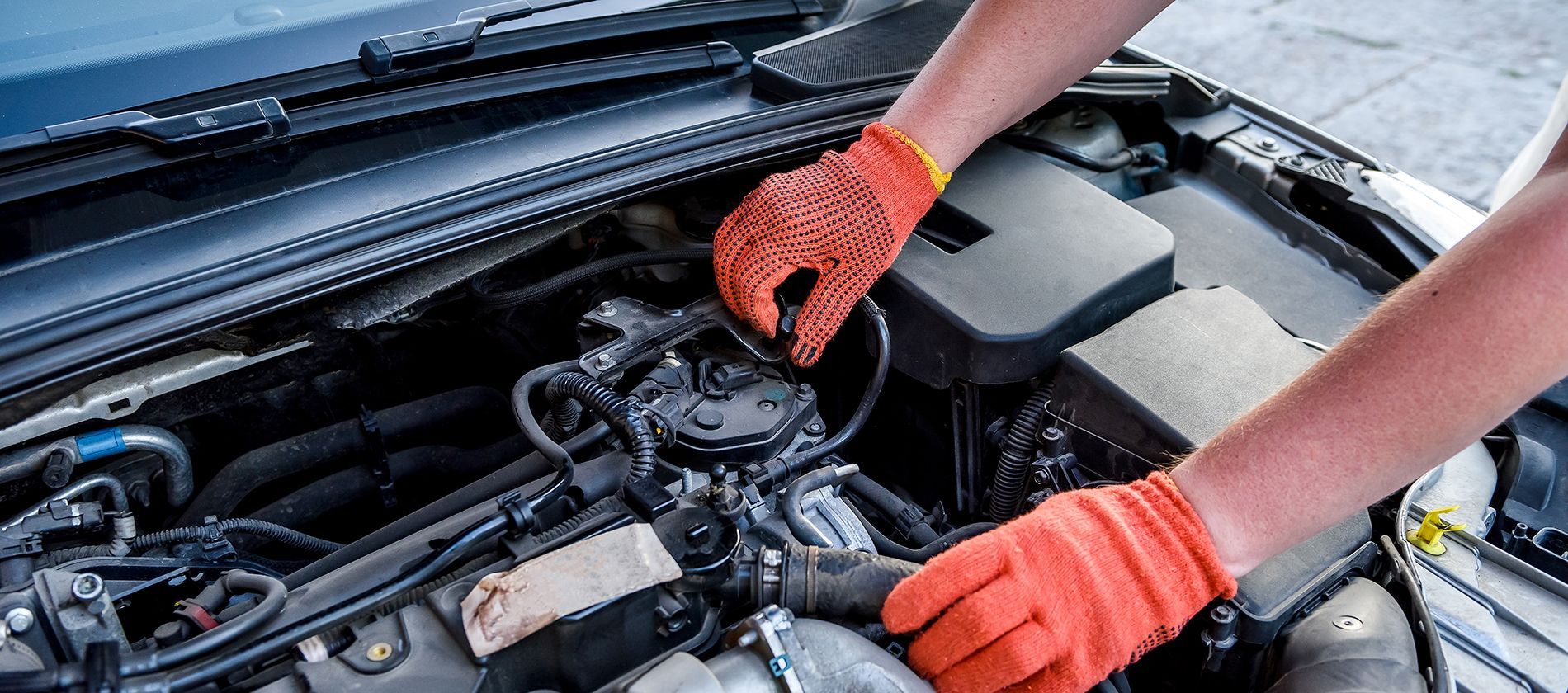 A person in orange gloves works on a car engine, inspecting parts under the hood.