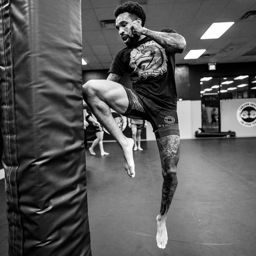 Martial artist in mid-air kick, tattooed leg. Training in a gym with others, black and white.
