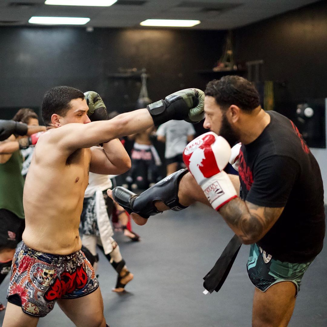 Two men practice Muay Thai in a gym. One throws a punch, the other blocks with a leg kick.