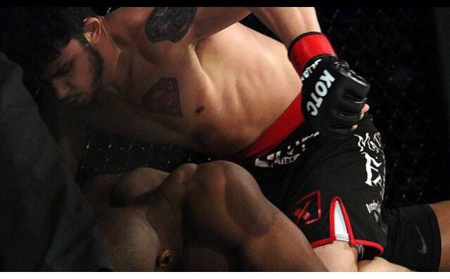 MMA fighter in black shorts with red trim, punching another fighter lying down in a cage.