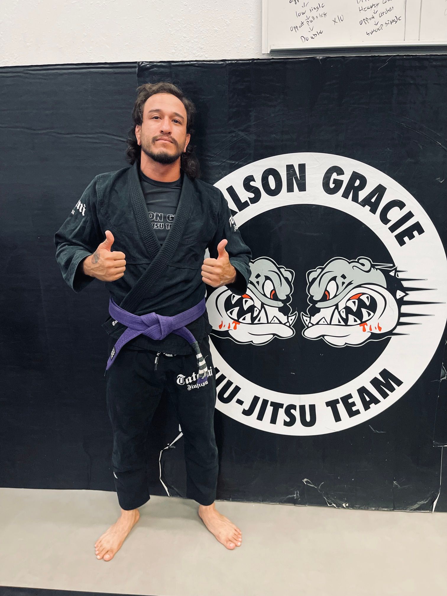 Man in a dark gi with a blue belt, standing in front of gym banners with skull and bulldog logos.