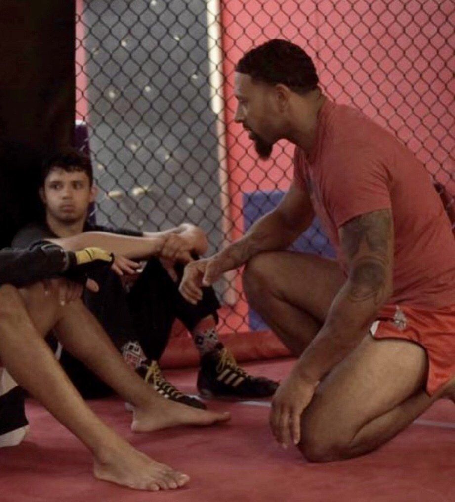 Three people in a gym: a man kneeling, another man sitting, and a partial view of a third's legs. The kneeling man is speaking.