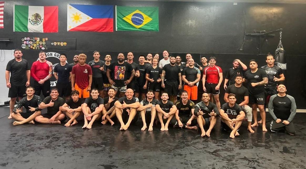 Group of people, mostly in black athletic wear, posing on a gym floor in front of flags.