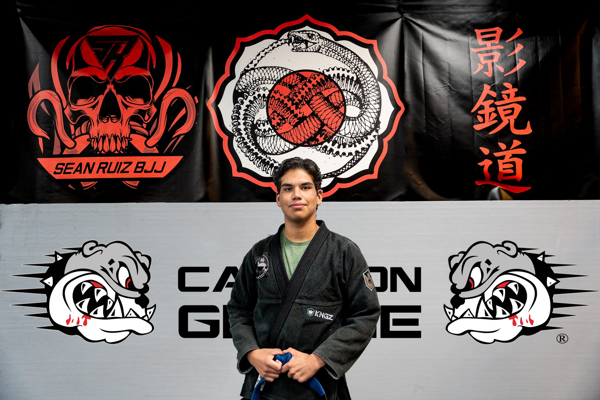 Man in a dark gi with a blue belt, standing in front of gym banners with skull and bulldog logos.