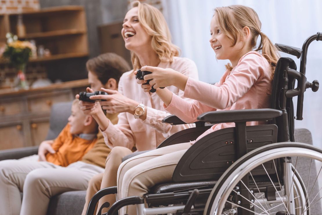 Due donne e un ragazzo ridono su un divano, una donna in sedia a rotelle, con in mano dei controller per videogiochi in un soggiorno.