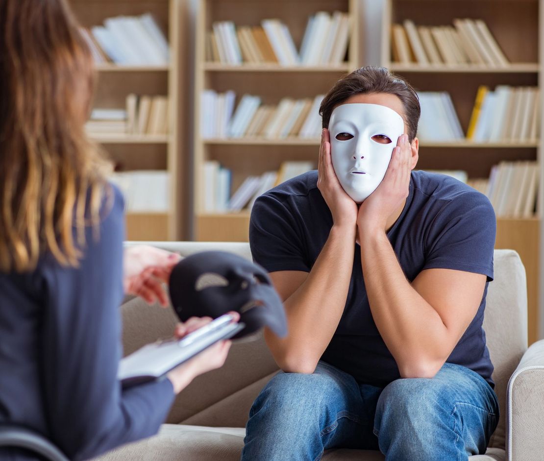 Una persona con indosso una mascherina bianca siede con la testa tra le mani durante una seduta di consulenza in una stanza con degli scaffali.