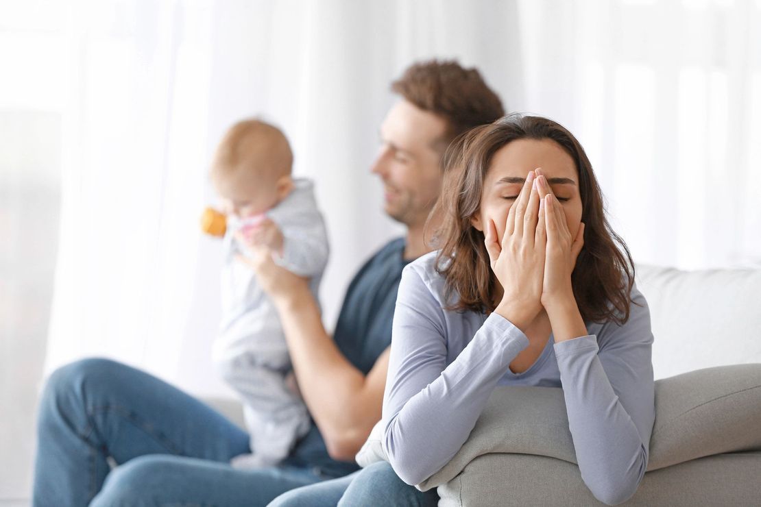 Una donna si copre il viso mentre è seduta su un divano con un uomo che tiene in braccio un bambino in un luminoso soggiorno.
