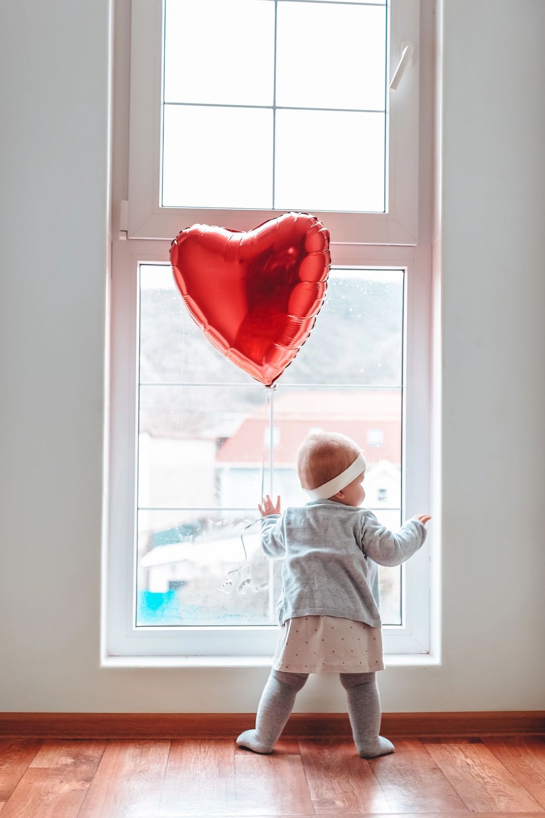 Un bambino vestito di bianco è in piedi accanto a una finestra e allunga la mano verso una decorazione a forma di cuore rosso.