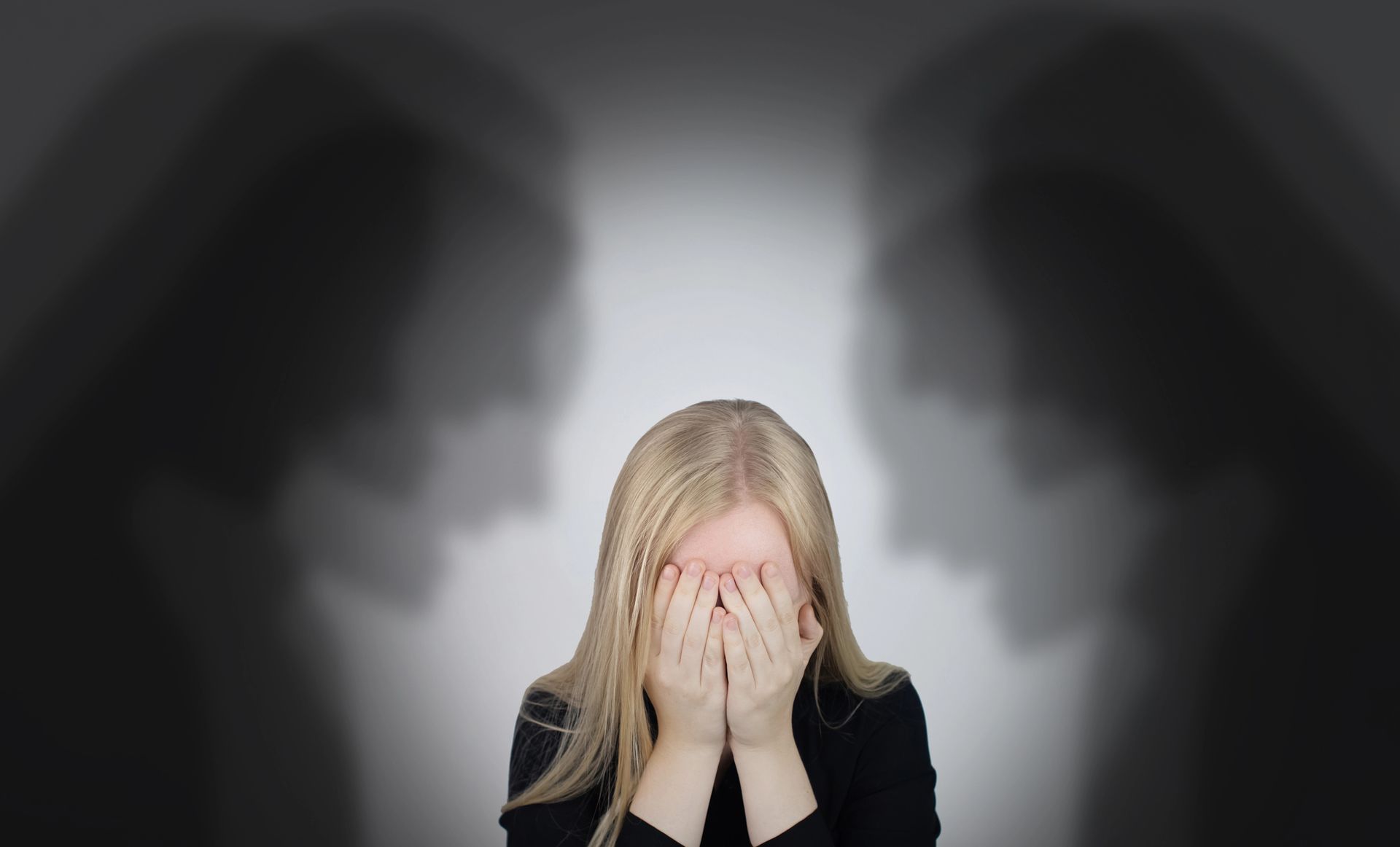 Una persona bionda si copre il viso con le mani, in piedi davanti a volti sfocati e in ombra su uno sfondo scuro.