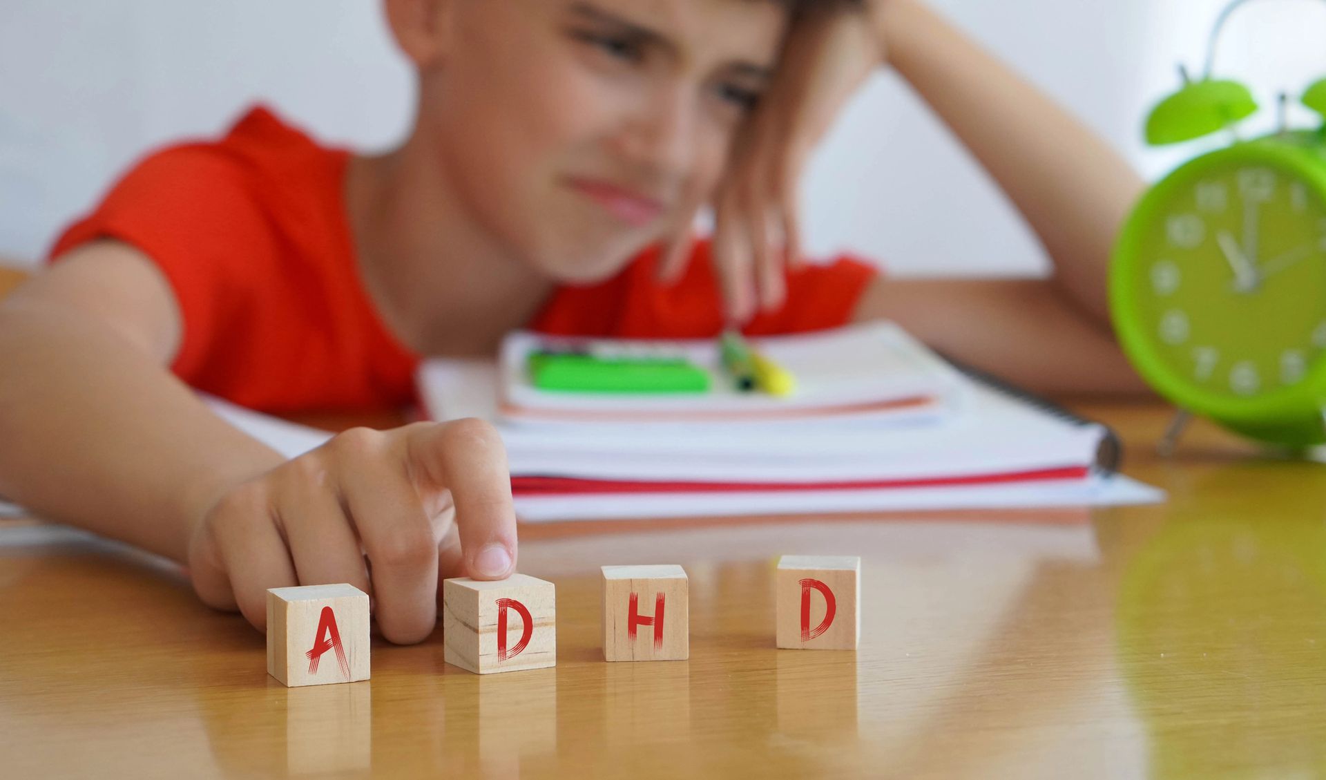Bambino con la maglietta rossa che gioca con i blocchi con le lettere a un tavolo, con un orologio verde e dei libri scolastici sullo sfondo.