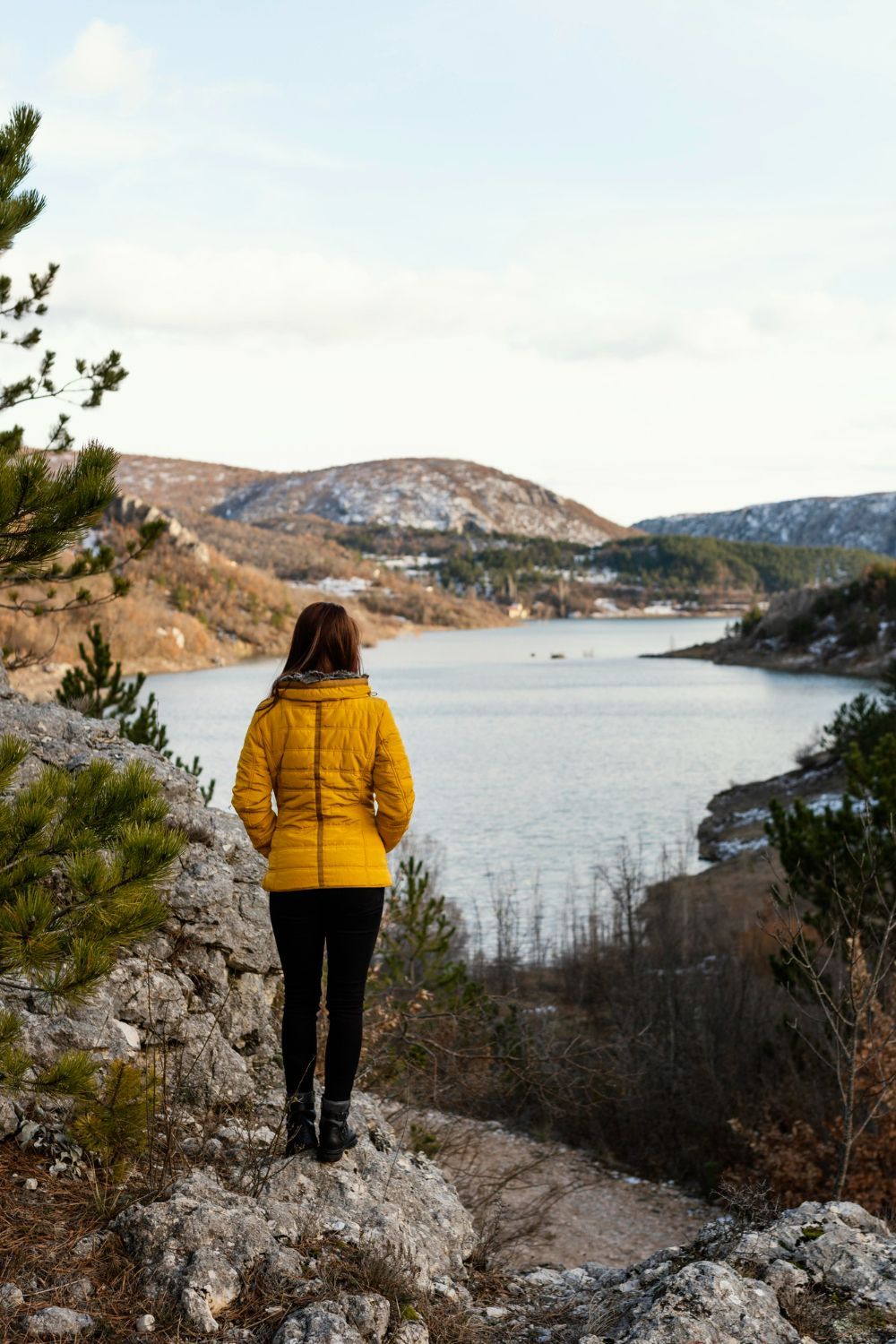A woman in a yellow jacket is standing on a rock overlooking a lake.