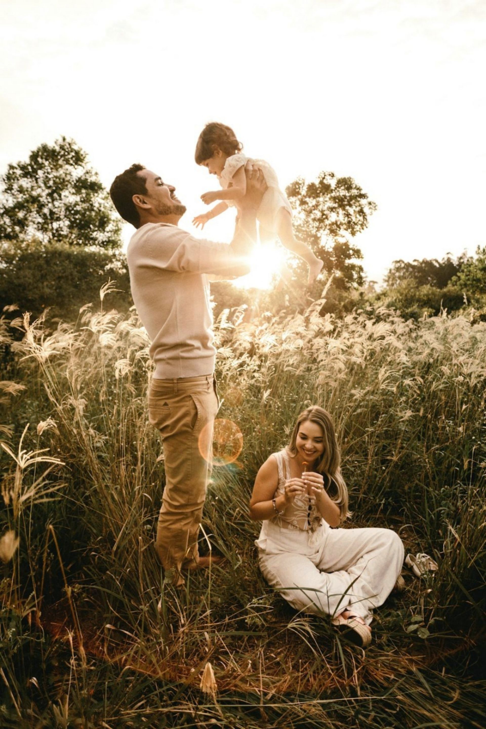 A man is holding a child in the air while a woman sits in the grass.