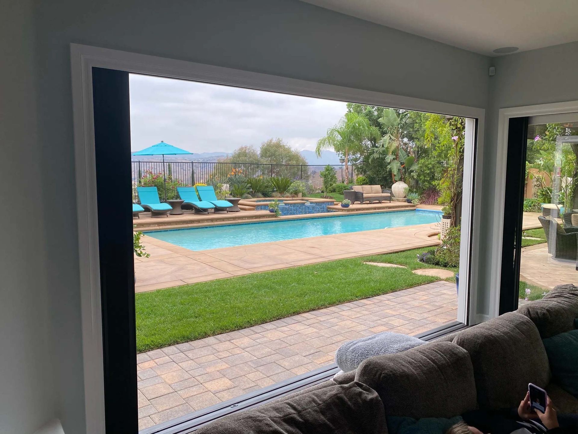 A man is sitting on a couch looking out a window at a swimming pool.