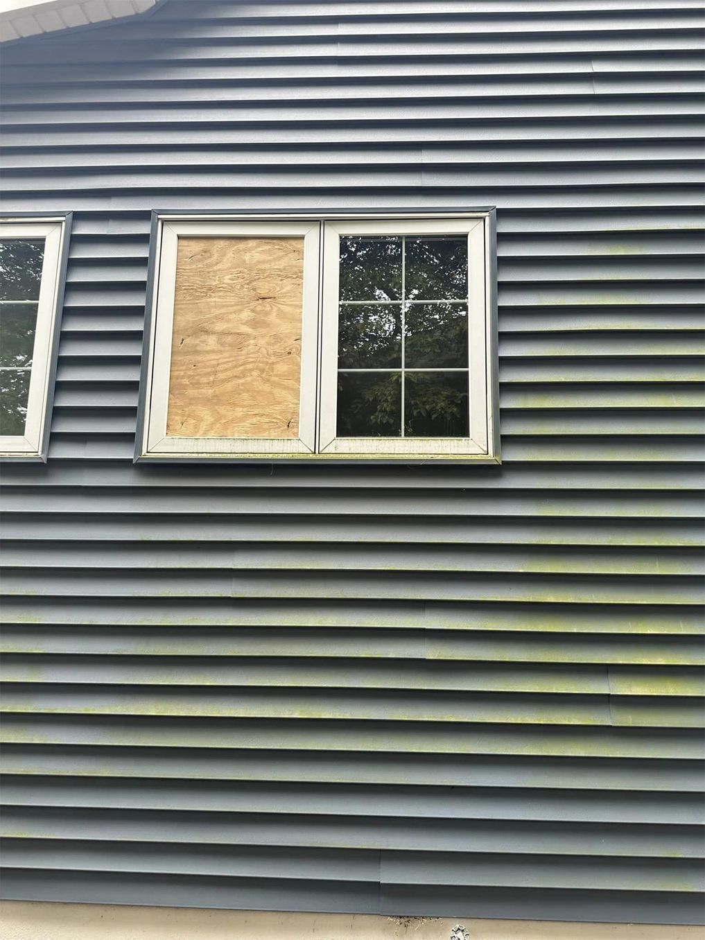 House siding with two windows, one boarded with plywood and one with white trim and glass panes.