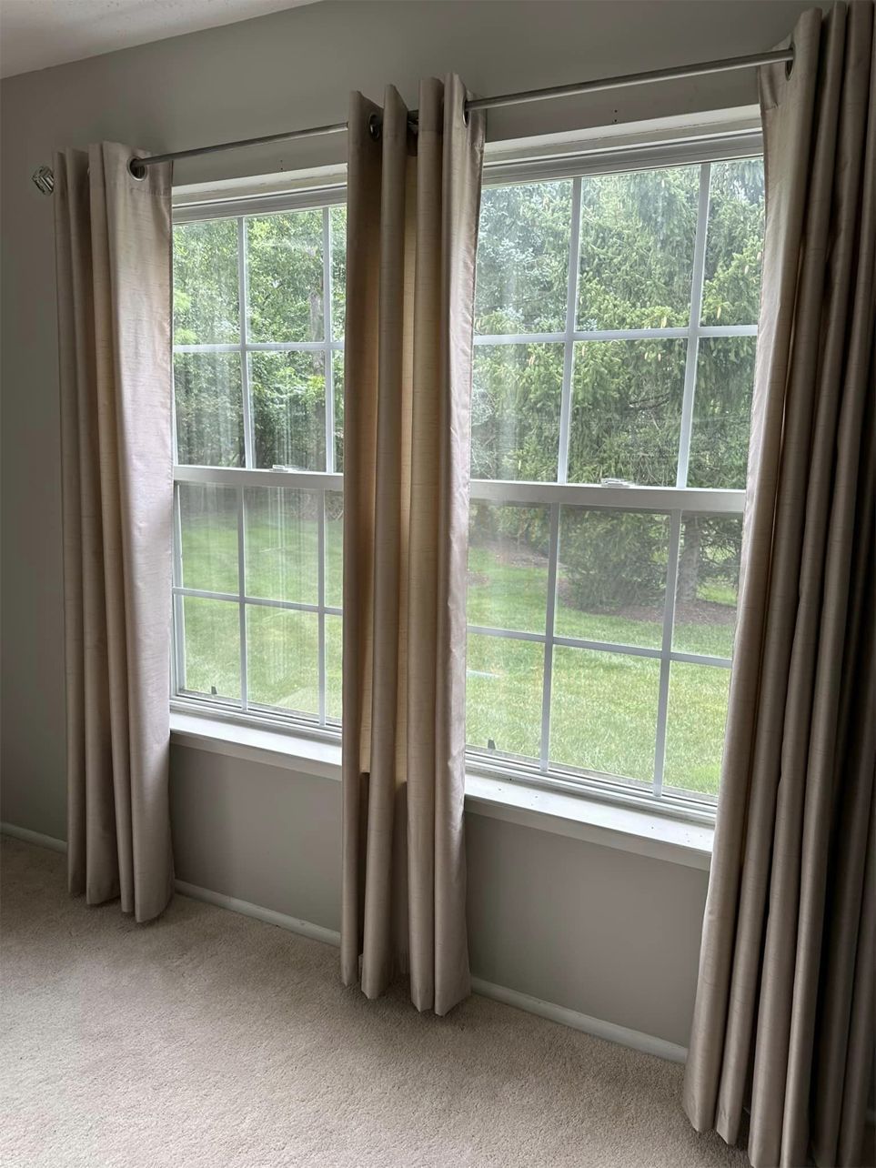 Empty room with two large windows, beige curtains, and a green yard outside