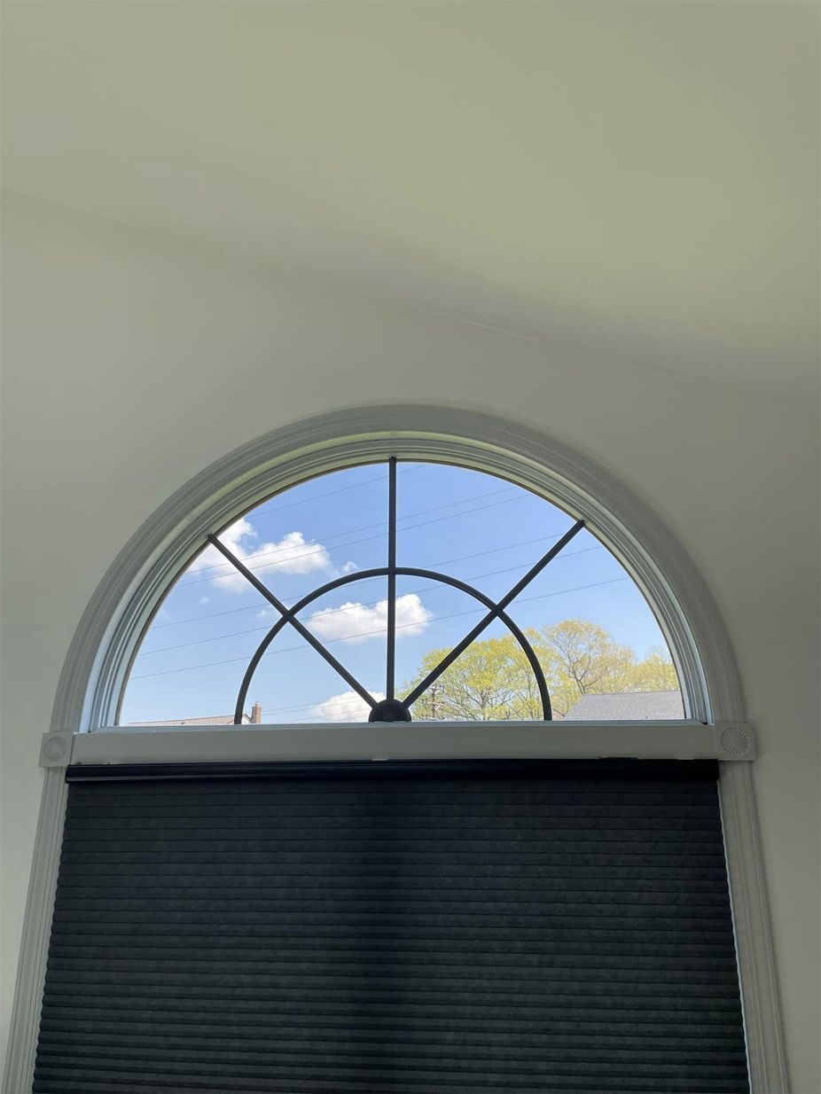 Arched window with decorative trim, partially covered by a dark roller shade, showing blue sky and trees outside