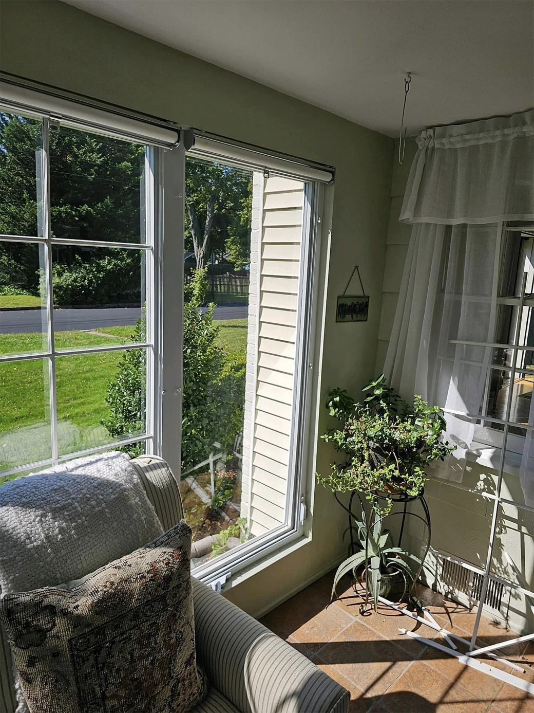 Bright sunlit living room corner with large windows, gray chair, potted plant, and a view of the lawn outside