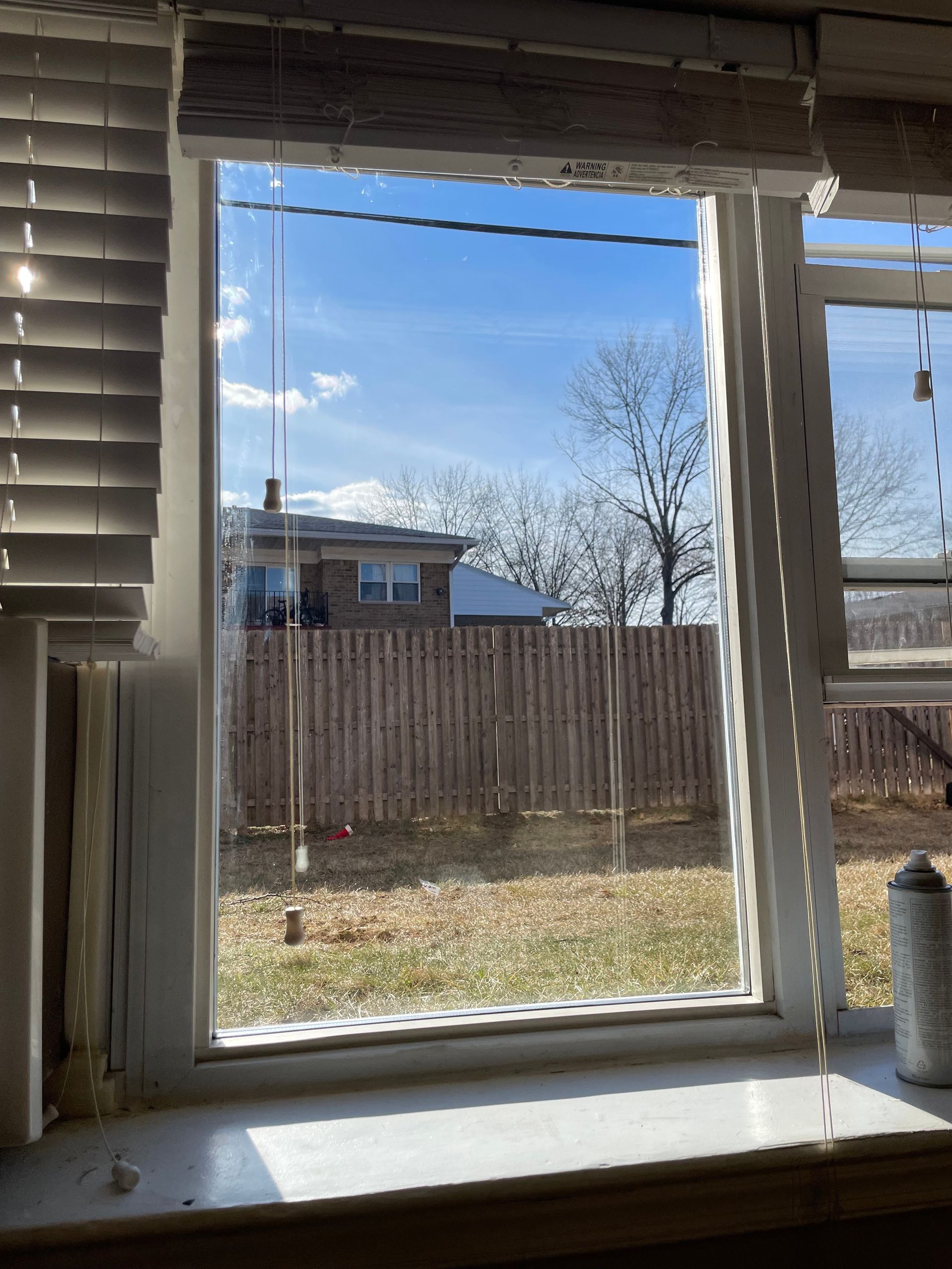 View through a window with blinds to a fenced backyard, house, and blue sky on a sunny day